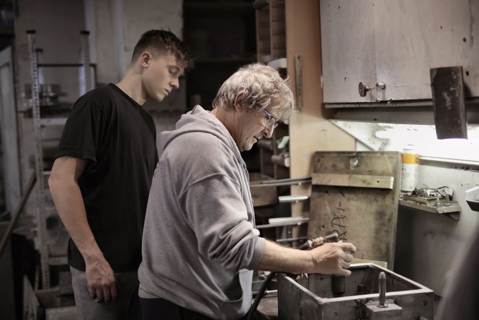 Side view of senior worker in eyeglasses and trainee handling detail on workbench in workshop